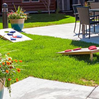A backyard scene featuring a cornhole game setup on a neatly manicured lawn, with adjacent patio furniture.