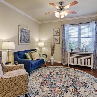 A cozy living room featuring a blue sofa, patterned armchair, round rug, and ample lighting from lamps and a ceiling fan.