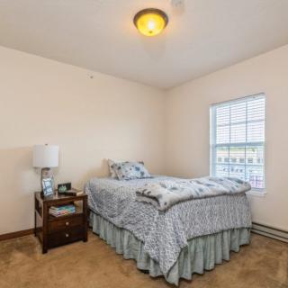 A cozy bedroom with a neatly made bed, nightstand, and a window letting in natural light.