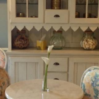 A cozy dining room with vintage furniture, featuring a table with a single flower in a vase and a classic cabinet in the background.