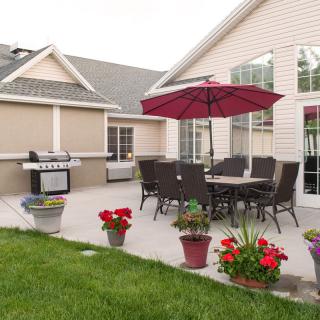 A cozy outdoor patio with a dining set, umbrella, and grill surrounded by potted plants.