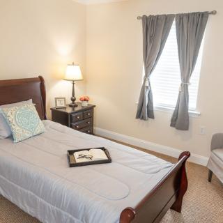 A cozy bedroom featuring a single bed with a nightstand and lamp, alongside a window with curtains.