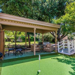 A cozy outdoor patio featuring a putting green and comfortable seating under a gazebo, surrounded by lush greenery.
