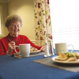 An elderly woman in a red sweater enjoying a meal at the dining table.