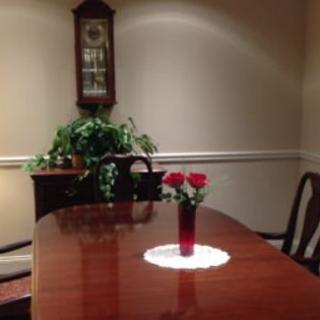 A dining room with a polished wooden table, adorned with a vase containing red flowers. A decorative clock and potted plant are in the background.