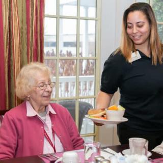 A caregiver serves a meal to an elderly woman in a dining setting.