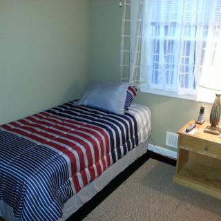 A cozy bedroom featuring a single bed with striped bedding, next to a nightstand with a lamp by the window.