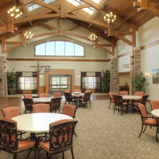 A spacious dining area with round tables, cushioned chairs, and wooden beams under a high ceiling with chandeliers.