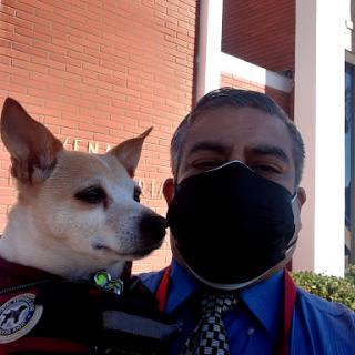 A person wearing a mask smiles while holding a dog outdoors.
