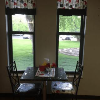 Cozy dining area with a table set for two by large windows overlooking a grassy outdoor scene.