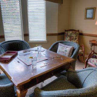 A cozy indoor room with a wooden table surrounded by chairs, featuring a lamp and large windows.