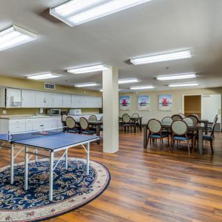 A spacious community recreation room featuring a ping pong table and dining area, illuminated by fluorescent lighting.