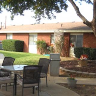 A serene outdoor patio area with a table and chairs, surrounded by greenery and adjacent to a low-rise brick building.