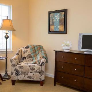 A cozy living room corner with a floral armchair, lamp, dresser, and television.