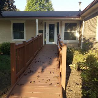 A house entrance with a wooden ramp leading to the front door, illuminated by sunlight.