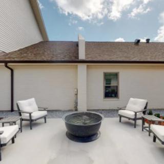 A cozy outdoor patio with cushioned chairs arranged around a fire pit, set against the backdrop of a house exterior under a partly cloudy sky.
