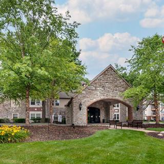 A stone building with an arched entrance surrounded by lush greenery and a well-maintained lawn under a partly cloudy sky.