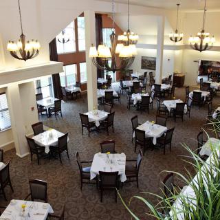 An elegant restaurant interior with neatly arranged, empty dining tables and chairs, featuring chandeliers and large windows.