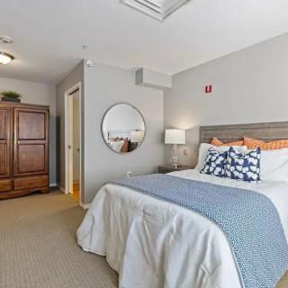 A cozy bedroom with a neatly made bed, featuring blue accents and decorative pillows. The room includes a wooden wardrobe, a round mirror, and a bedside lamp.