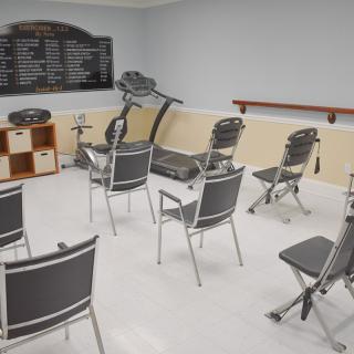 An indoor classroom setup with chairs arranged in a semi-circle and exercise equipment in the background.