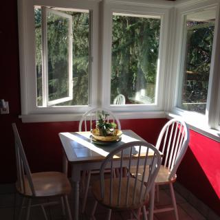 A cozy kitchen nook with a wooden table and chairs, bathed in sunlight from large windows.