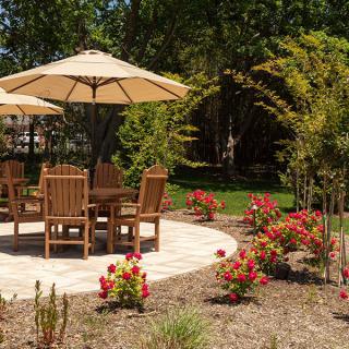 A garden patio area featuring wooden chairs and a table under an umbrella, surrounded by vibrant flowers.