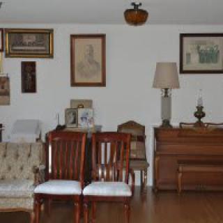 A cozy living room featuring a sofa, wooden chairs, a piano, and various framed artworks on the wall.