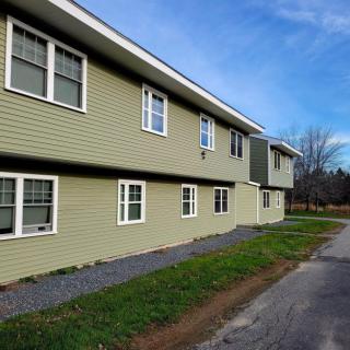 A residential building with green siding and multiple windows, situated next to a road under a clear blue sky.