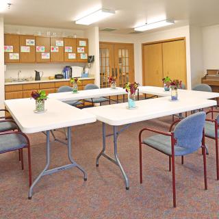 A meeting room with tables arranged in a U-shape, surrounded by chairs. The space includes a kitchenette and decorative elements.