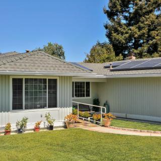 A single-story house with solar panels on the roof, surrounded by a well-maintained lawn and trees under a clear blue sky.