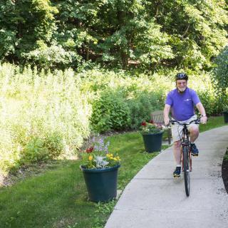 A person cycling on a paved pathway surrounded by lush greenery.