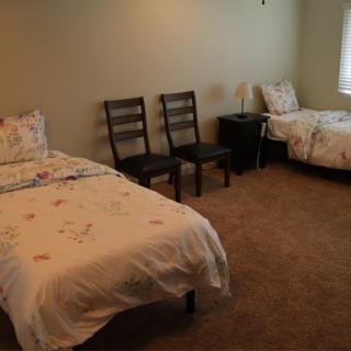 A simple bedroom featuring two beds, a nightstand, and chairs on a carpeted floor.