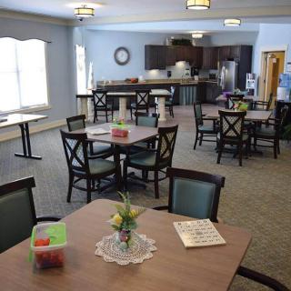A community dining area with multiple tables and chairs, featuring a kitchen in the background.