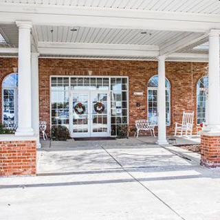 A building entrance featuring double doors with wreaths, flanked by columns and rocking chairs on a brick patio.