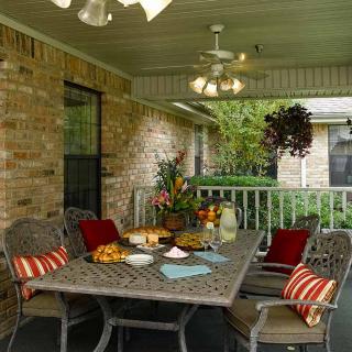 A cozy covered patio with a dining table set, decorated with cushions and surrounded by greenery.