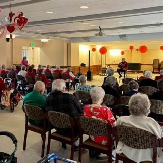 A group of seniors seated in a room decorated with red balloons and hearts, attending an event.