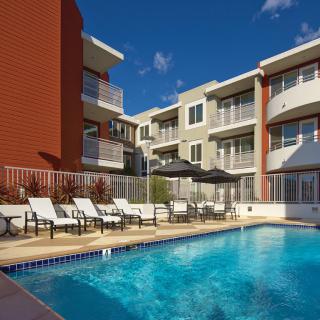 A modern apartment complex with a pool and lounge chairs under a blue sky.