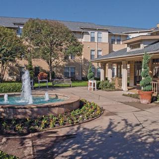 A picturesque courtyard with a central fountain, surrounded by a garden and buildings under a clear blue sky.