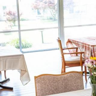 A bright dining room with tables and chairs arranged near large windows, allowing natural light to flood in. A vase of flowers sits on one of the tables.