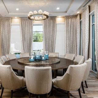 A modern dining room featuring a large round table surrounded by upholstered chairs, with elegant curtains and a decorative chandelier overhead.