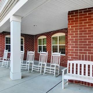A covered porch with white rocking chairs and a bench against a brick wall.