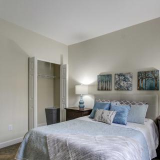 A cozy, well-decorated bedroom featuring a bed with blue and white bedding, a nightstand with a lamp, artwork on the wall, and an open closet.