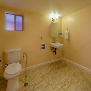 An accessible bathroom with a toilet, sink, and mirror, featuring a handrail and tiled floor.