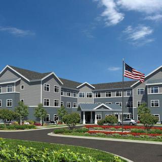 A large residential building with an American flag in front, surrounded by landscaping, under a blue sky with clouds.