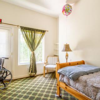 A cozy bedroom with a bed, wheelchair, and natural light coming through large windows.