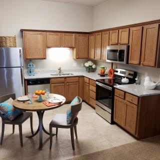 A modern kitchen with wood cabinets and stainless steel appliances, featuring a small dining table with chairs.