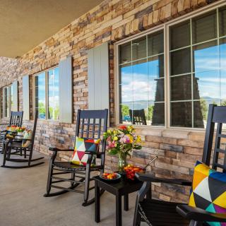 A cozy porch featuring rocking chairs with colorful pillows, a brick exterior, and decorative flowers on small tables.
