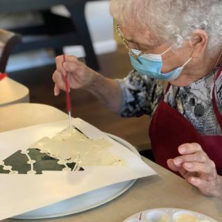 An elderly woman wearing a mask is painting using a stencil at a table.