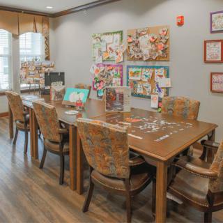 A cozy dining area with a large wooden table and upholstered chairs, adorned with colorful artwork and natural light from the windows.