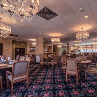 A well-lit restaurant dining room featuring elegant chandeliers, wooden tables, and upholstered chairs on a patterned carpet.